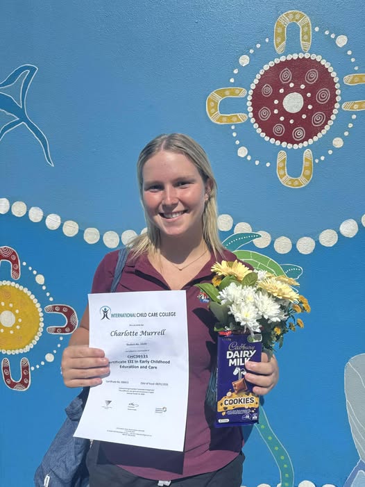 Tillys Waratah A smiling person holds a certificate, flowers, and a chocolate bar, standing against a colorful mural featuring abstract patterns and symbols.