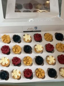A box of 24 mini cupcakes with red, yellow, navy, and white frosting arranged in rows. The cupcakes are neatly displayed in a white box with a clear lid. The colors and arrangement create a festive and inviting appearance.