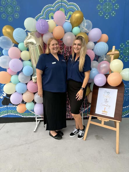Tillys Cooranbong Two smiling women in blue shirts stand in front of a colorful balloon arch with gold and pastel balloons. A sign on an easel reads "Happy 1st Birthday."