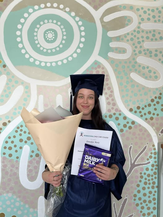 Tillys Rutherford A person in a graduation cap and gown smiles, holding a bouquet, certificate, and a chocolate bar. The background features a colorful, abstract mural.
