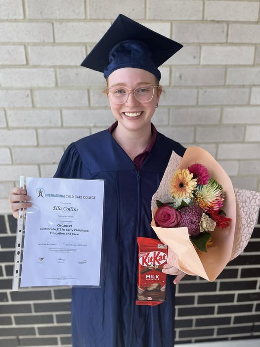 A person stands in front of a brick wall wearing a graduation gown, cap, and glasses. She is smiling, while holding a certificate, chocolate and bunch of flowers.