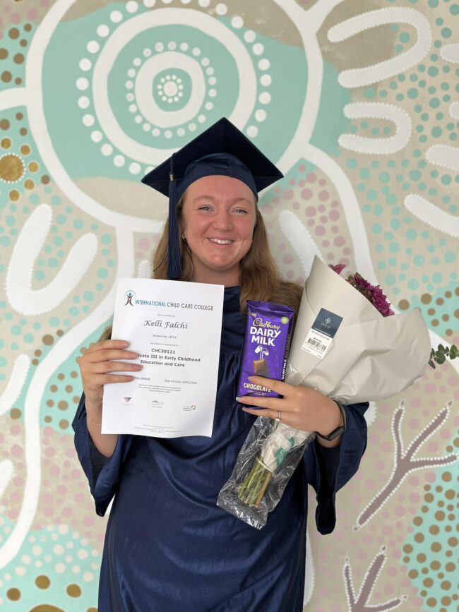 a person stands infront of a colourful mural wall, holding chocolate, a bunch of flowers and a certificate. They are wearing a graduation gown and cap.