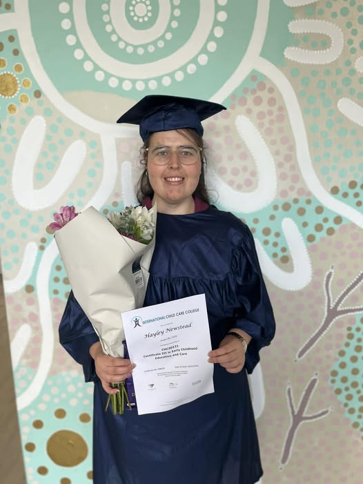 A smiling person stands infront of a colourful mural on a wall, wearing a graduation gown and cap. She is holding a certificate and a bunch of flowers.