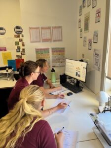 Tillys Fern Bay Three people look at a computer screen within an office space. They are holding pens and writing notes on a piece of paper each. The walls have posters on them, there is stationary and office equipment on the desk.