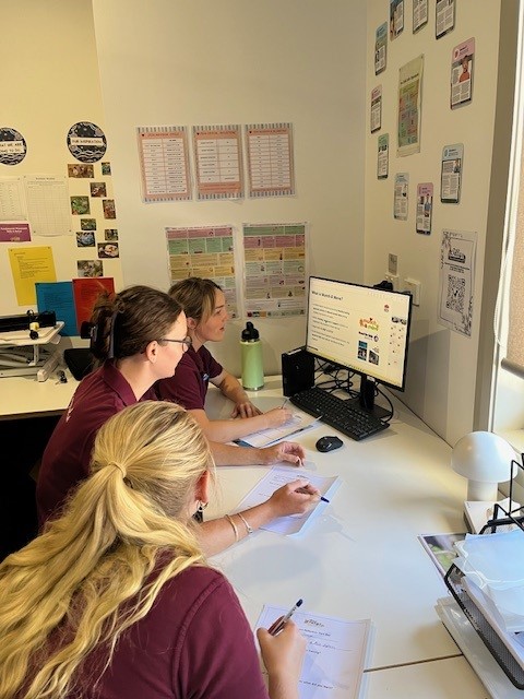 Three people look at a computer screen within an office space. They are holding pens and writing notes on a piece of paper each. The walls have posters on them, there is stationary and office equipment on the desk.