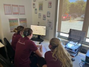 Tillys Fern Bay Three people sit in an office space facing toward a computer. There is posters on the wall, office equipment and stationary.
