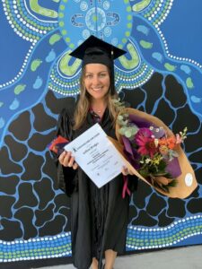 A smiling lady stands in front of a blue painted mural wearing a black graduation gown and cap. She is holding a certificate, a bunch of flowers and a block of chocolate.