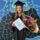 A smiling lady stands in front of a blue painted mural wearing a black graduation gown and cap. She is holding a certificate, a bunch of flowers and a block of chocolate.